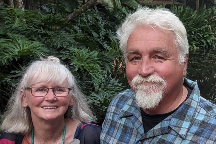 A woman with grey hair and glasses stands beside a man with grey hair and beard wearing a blue checked shirt