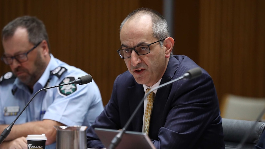 Michael Pezzullo looks towards senators out of shot as he gives evidence with an AFP officer sitting next to him