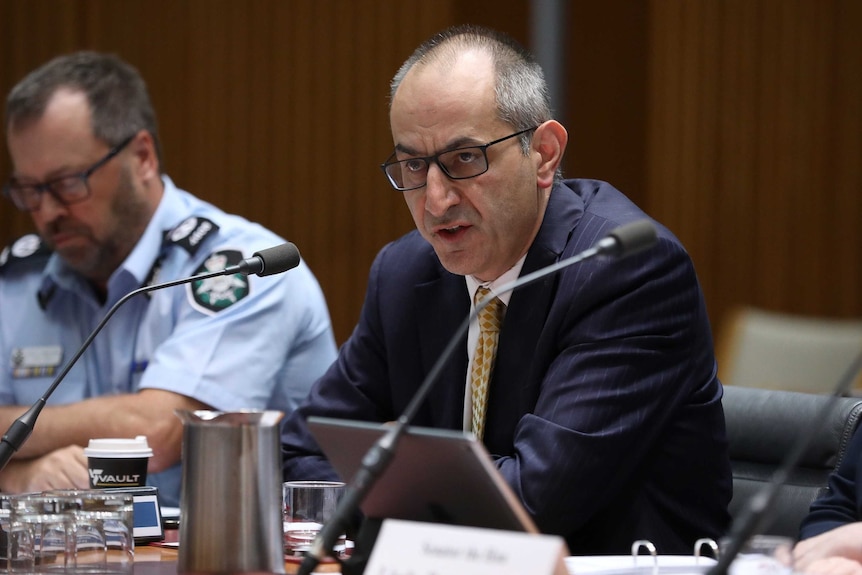 Michael Pezzullo looks towards senators out of shot as he gives evidence with an AFP officer sitting next to him.