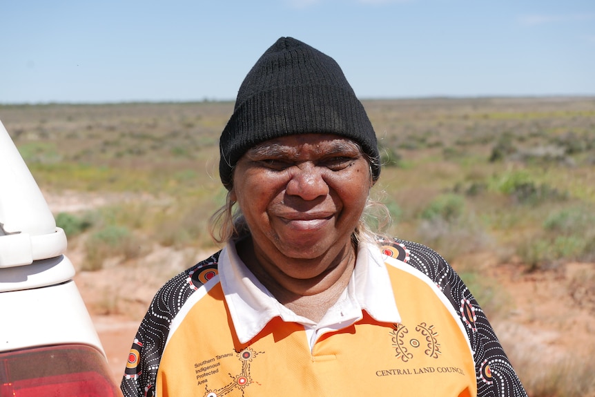 An Aboriginal woman in the desert wears a black beanie and squints in the sunlight, leaning against a white car.