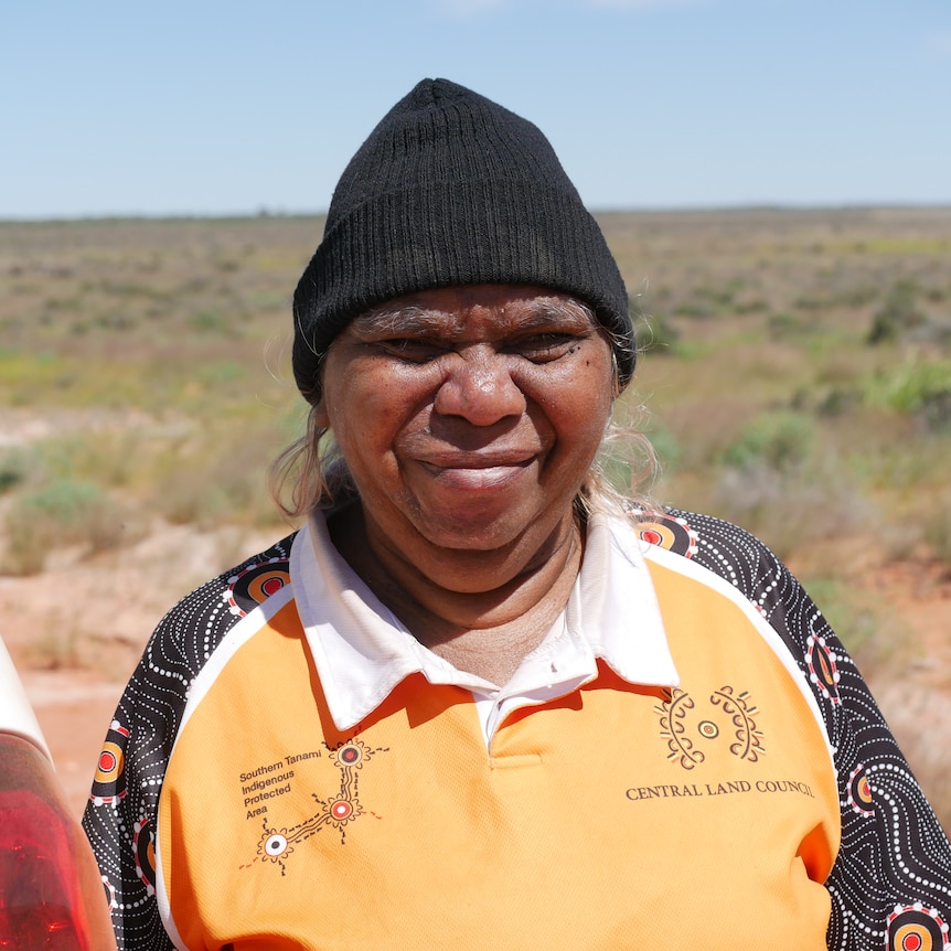 An Aboriginal woman in the desert wears a black beanie and squints in the sunlight, leaning against a white car.