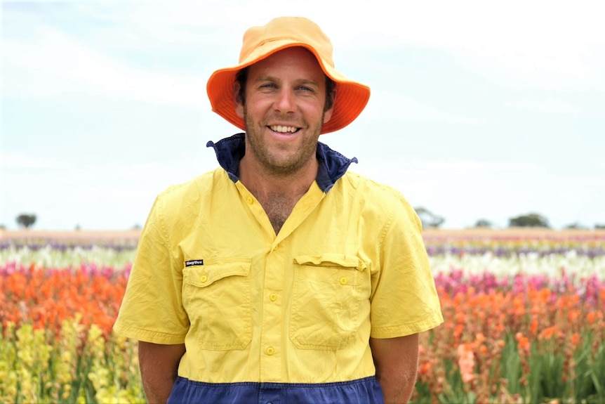 A middle aged man with a fluro orange bucket hat and bright high vis shirt smiles in a field of colourful flowers 