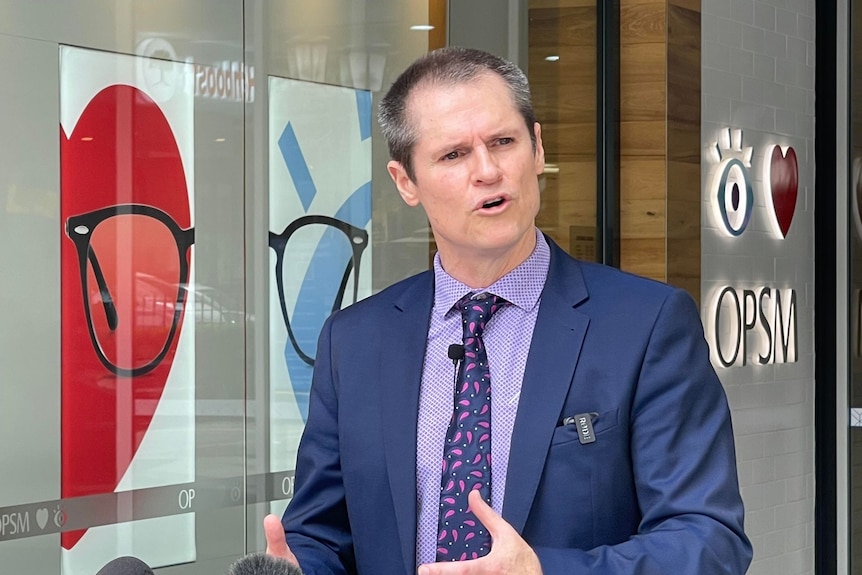 A dark-haired man in a dark suit stands outside a shopfront and speaks to the media.