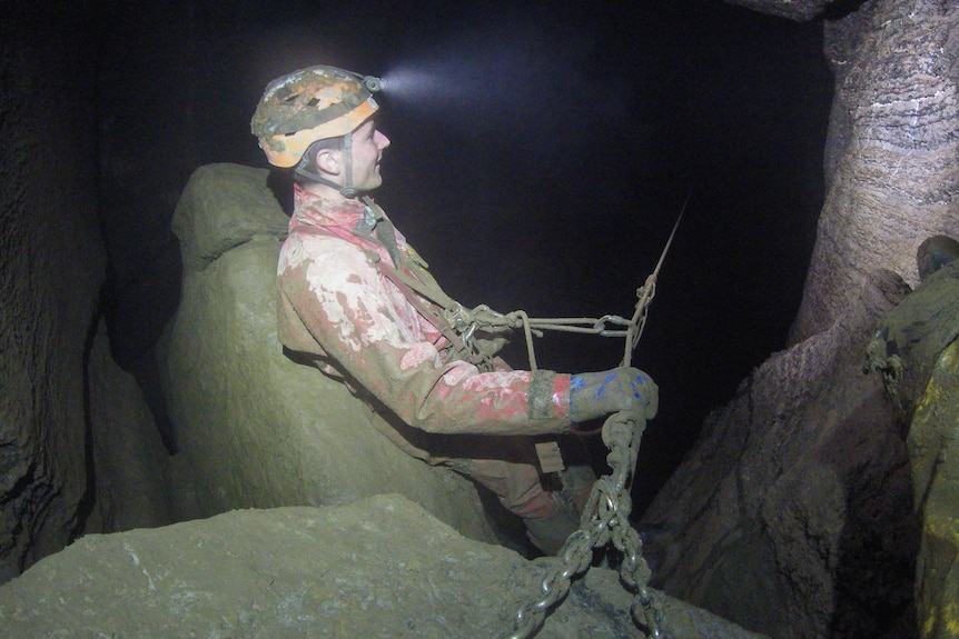 A man with a helmet, head torch and muddy overalls at the end of a cable in a dark cave