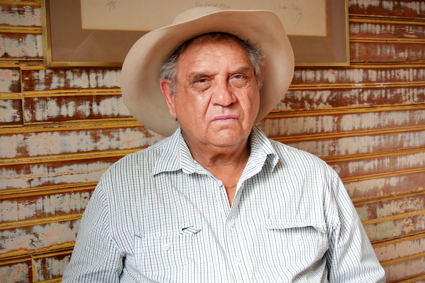 a man with an akubra-style hat sits against a brown wall, looking at the camera with a serious expression