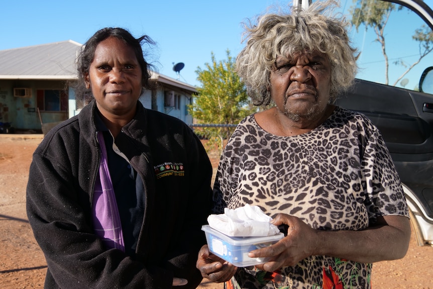 A young Indigenous worker in uniform next to an elderly Indigenous woman holding a transparent lunchbox outside an outback house