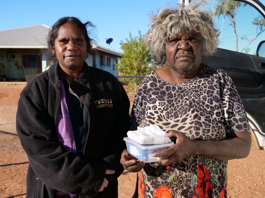 A young Indigenous worker in uniform next to an elderly Indigenous woman holding a transparent lunchbox outside an outback house