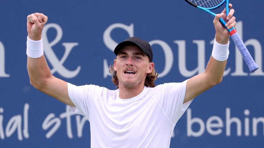 A cap-wearing Australian tennis player smiles and holds his racquet in the air in celebration after a win.