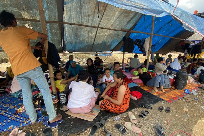 Dozens of people under a shelter made of bamboo and tarps. 