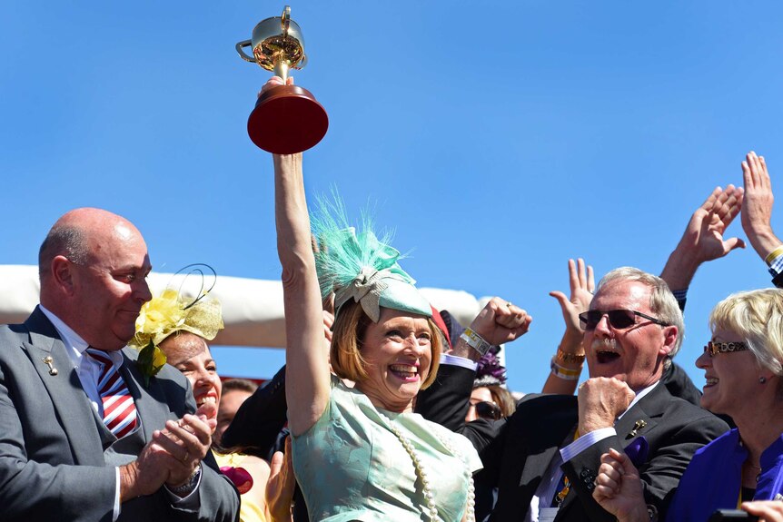 Woman in a green dress surrounded by men holds the Melbourne Cup aloft
