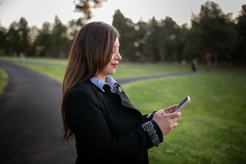 A woman stands looking at her mobile phone in a park
