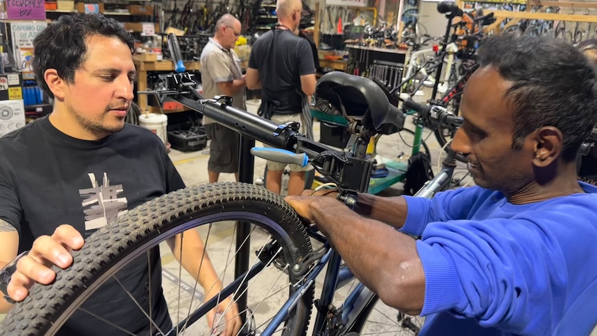 two men fix the rear wheel of a bike in a workshop with two men talking in teh background