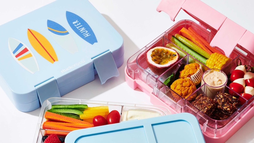 Pink and blue lunch boxes filled with chopped vegetables, passionfruit, berries, yoghurt, baby bocconcini, and home-baked treats
