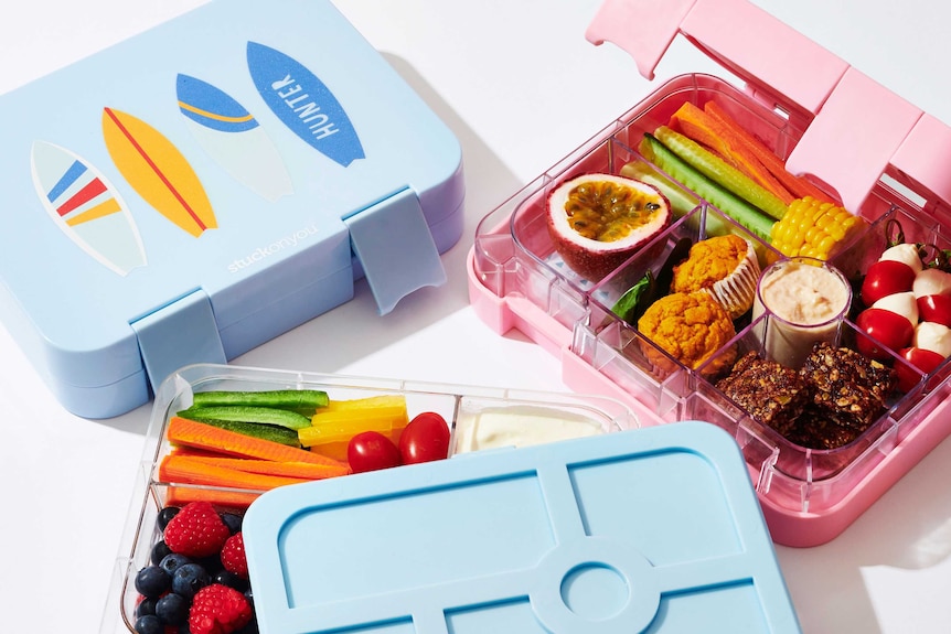 Pink and blue lunch boxes filled with chopped vegetables, passionfruit, berries, yoghurt, baby bocconcini, and home-baked treats
