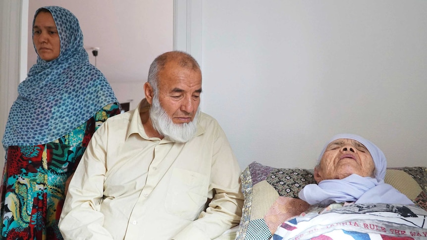 An elderly woman in a light blue head scarf lies in bed beside a bearded man and another woman in traditional Afghan dress.