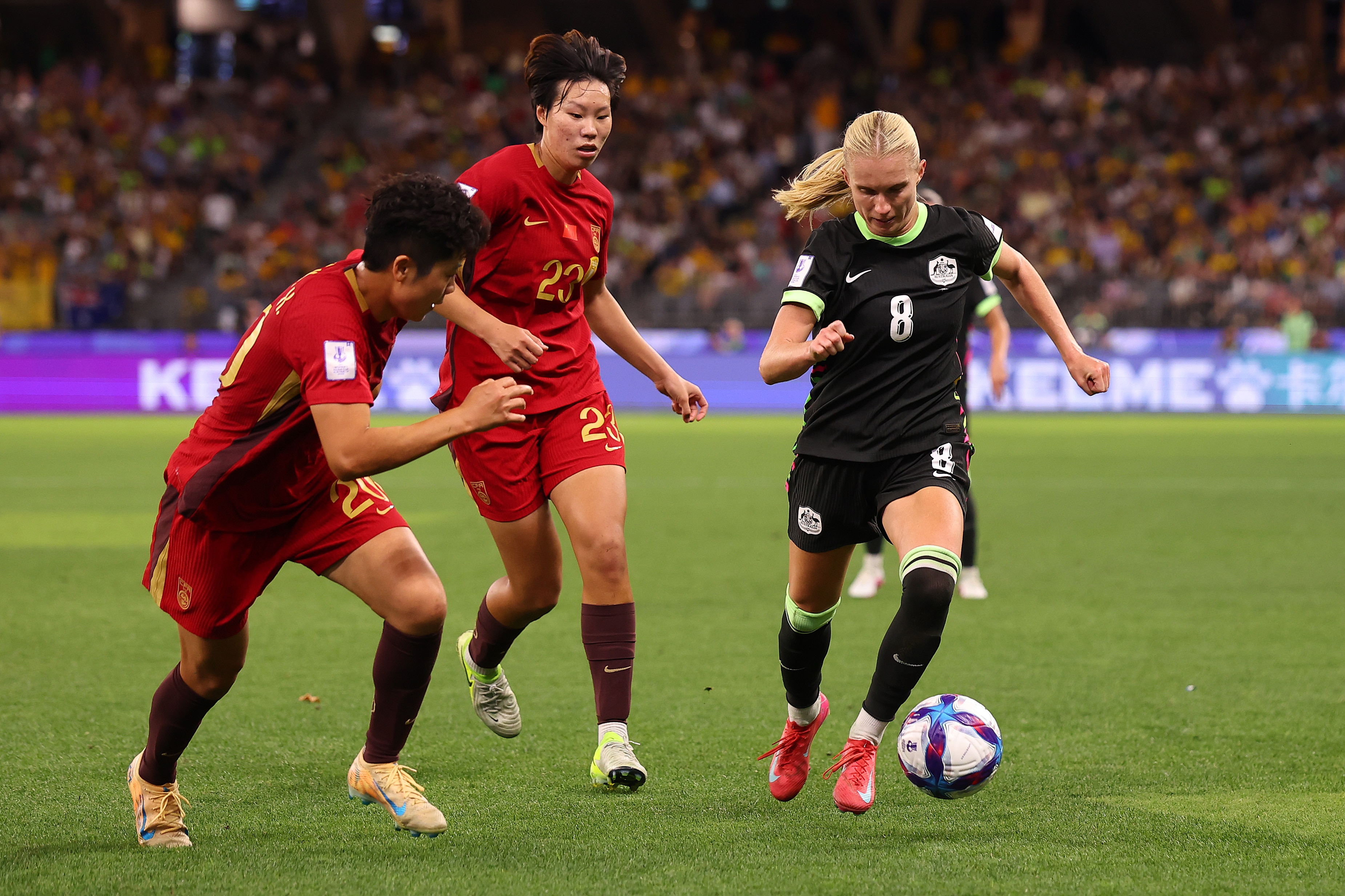 Uma jogadora de futebol de verde corre com a bola, jogadores de vermelho ao lado dela