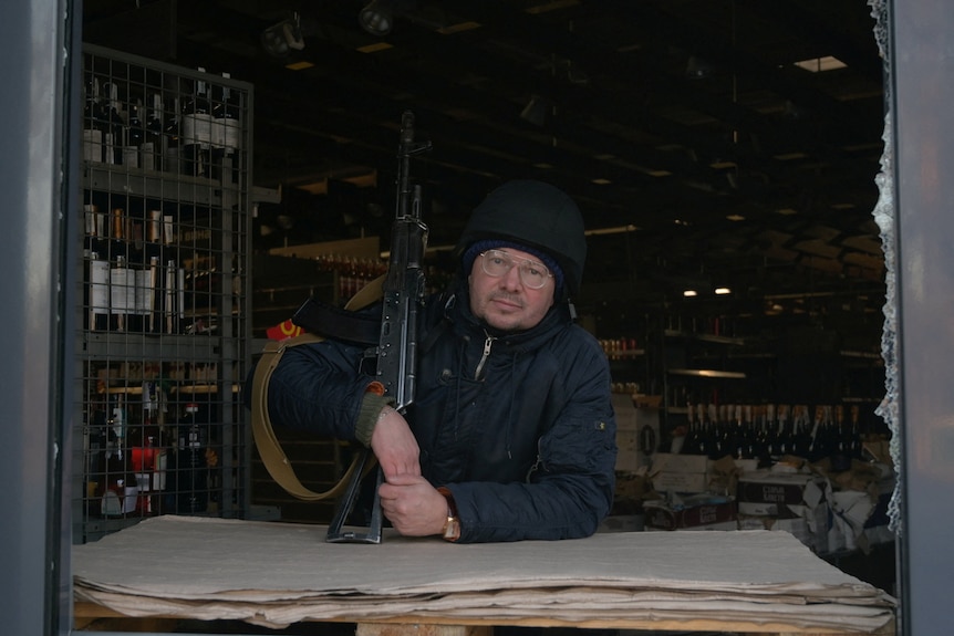 A man with a Kalashnikov rifle stands in the window of a bottleshop looking at the camera.