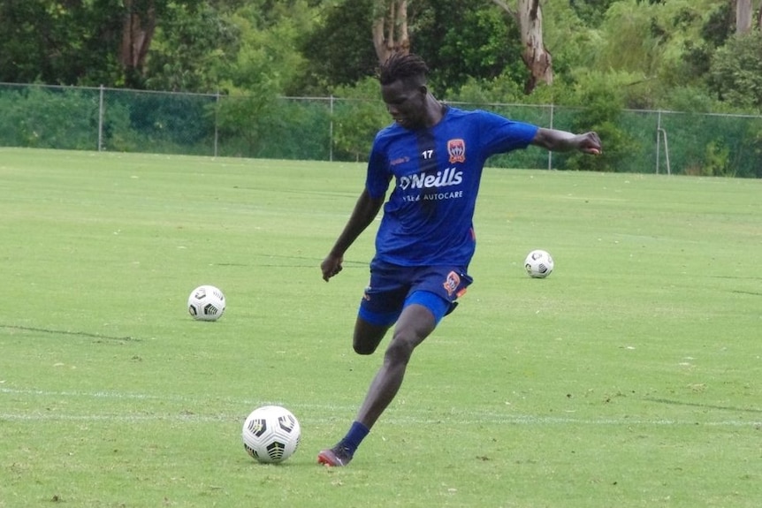 An African man in blue sports top and shorts prepares to kick a soccer ball while practising on a  green sports field.