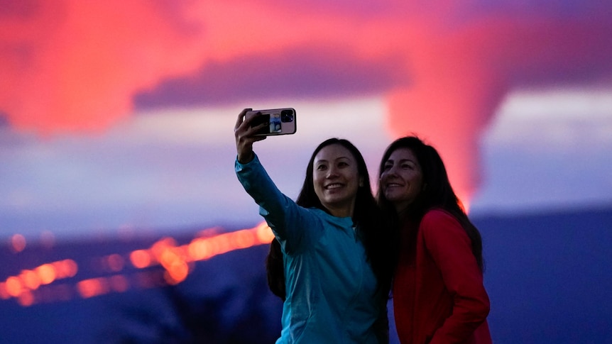 Two people take a selfie in front of an erupting volcano.