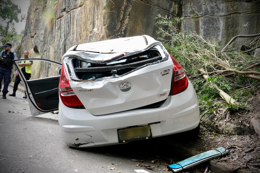 A damaged white car rests on a road at the bottom of a cliff with police looking on