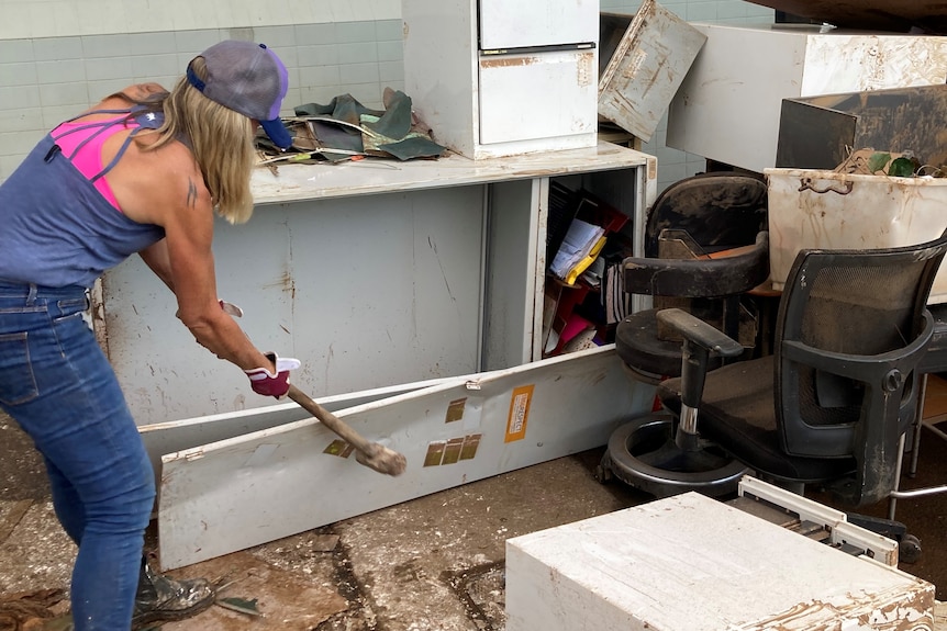 A woman in working gear takes a sledgehammer to a flooded out cabinet