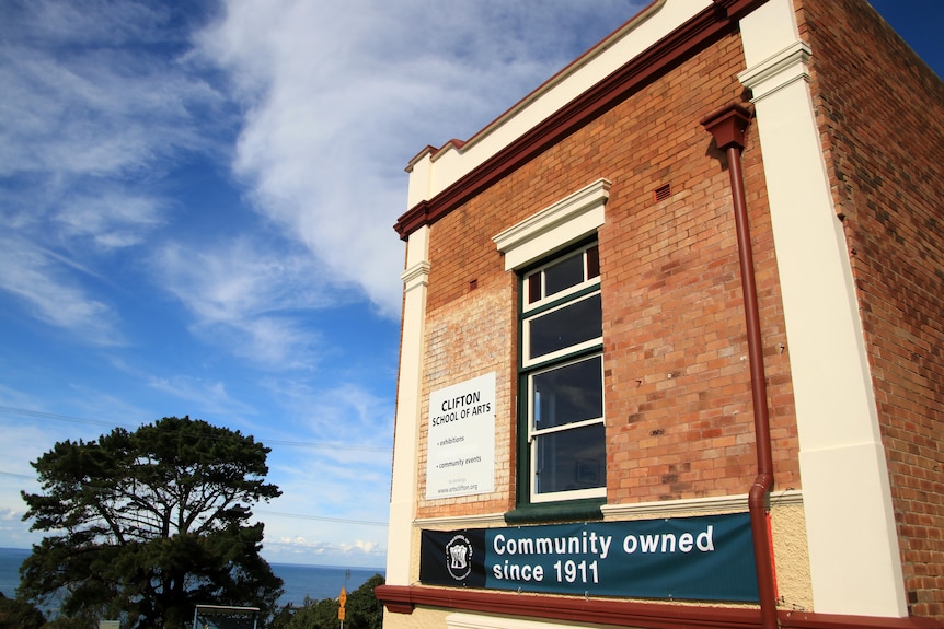 The brick side of the Clifton School of Arts building with a sign promoting it is community owned and blue sky behind it.