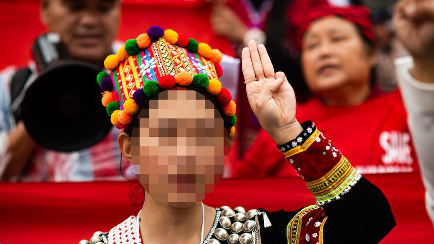 A woman in colourful traditional Kachin dress gives a three-finger salute. Her face is blurred.