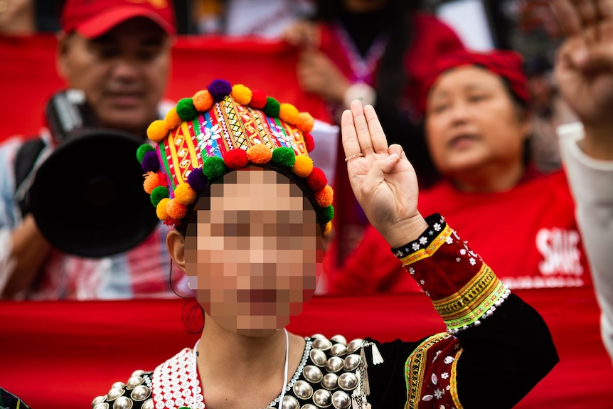 A woman in colourful traditional Kachin dress gives a three-finger salute. Her face is blurred.