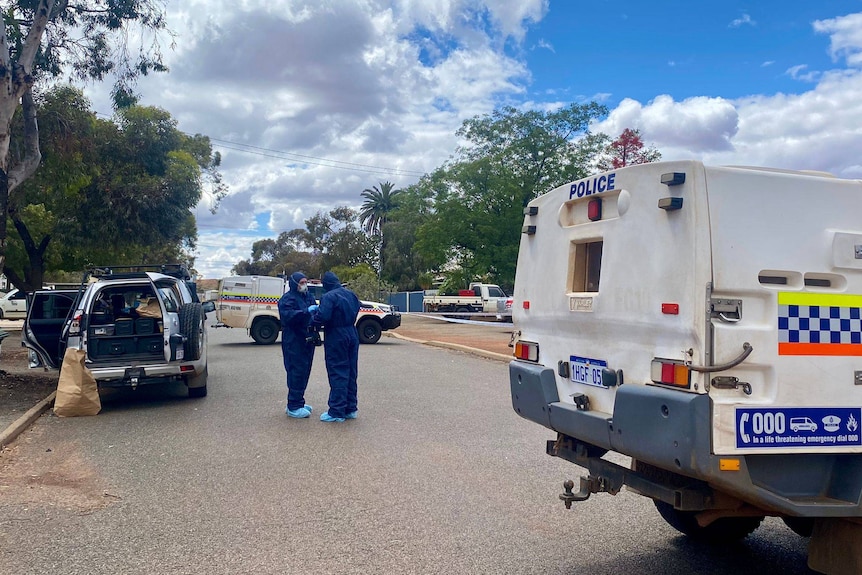 Two men in protective suits stand in the middle of a street, with police vehicles on the left and right.