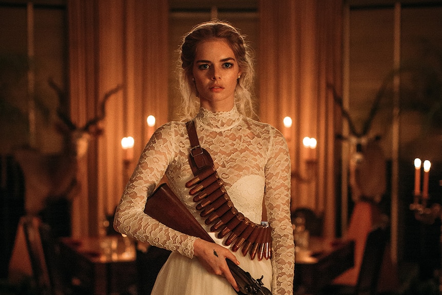 Samara Weaving stands in candlelit room wearing lace wedding dress with cartridge belt slung over shoulder and holding shotgun.