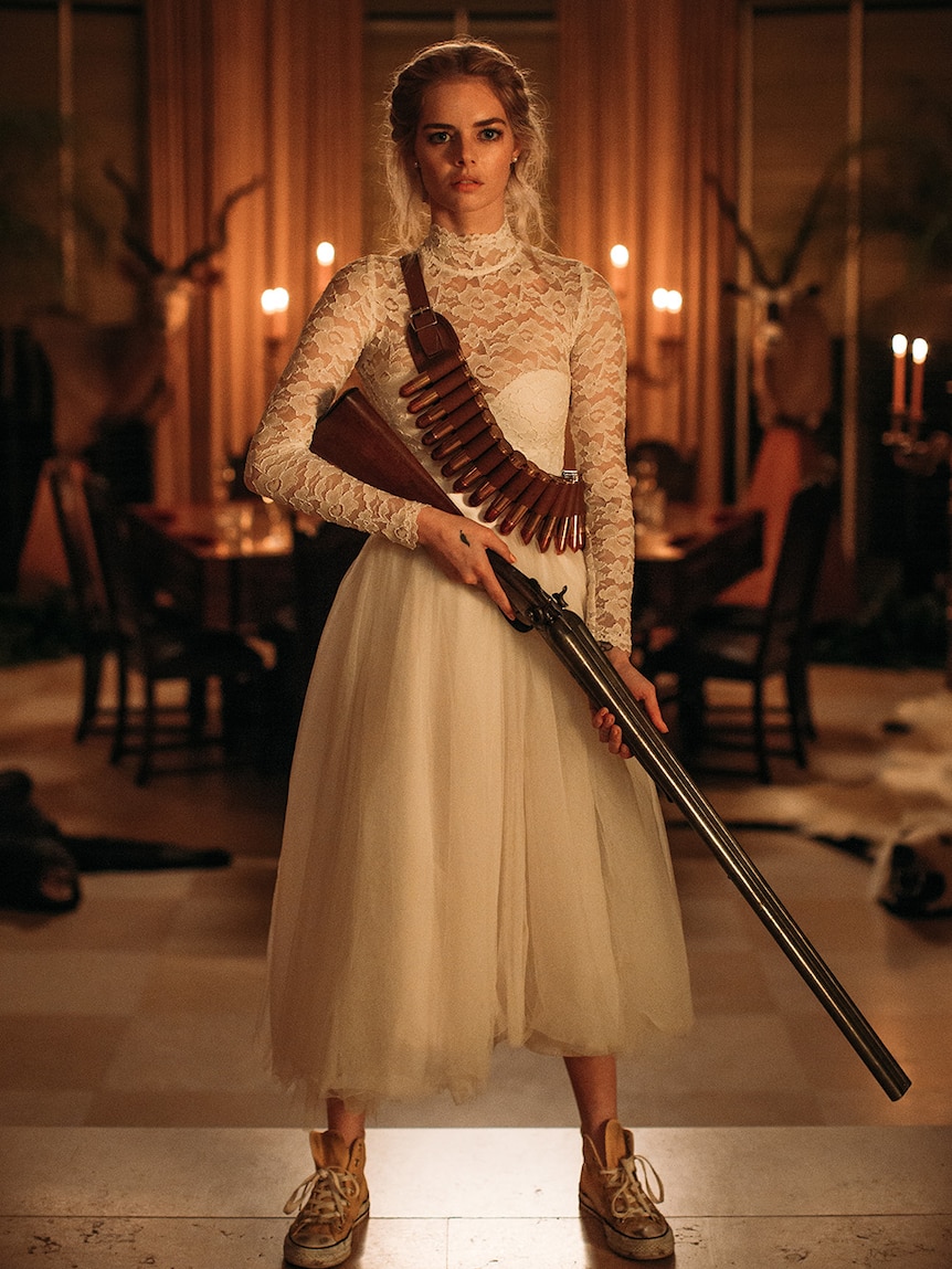 Samara Weaving stands in candlelit room wearing lace wedding dress with cartridge belt slung over shoulder and holding shotgun.