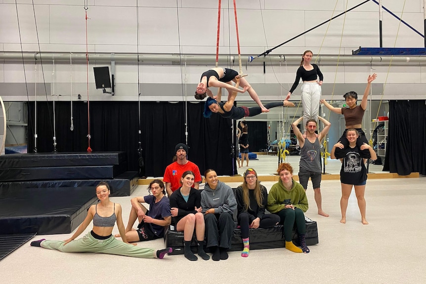 A group of circus arts students in a gym, some sitting on the floor, two hanging on ropes and two on others' shoulders