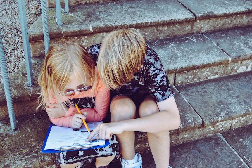 A bird's eye view of two young blonde children crouching over a clipboard and sitting on a cement step outside.