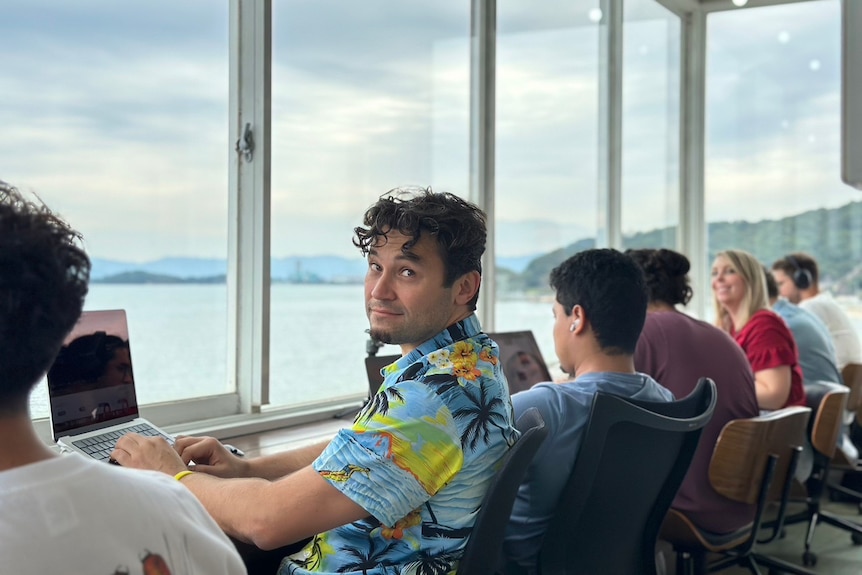 A man in a Hawaiin top looks over his shoulder at the camera as he sits in a row of folk working on laptops with