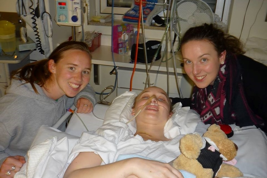 Woman lying in hospital bed with tubes and two woman by her side.