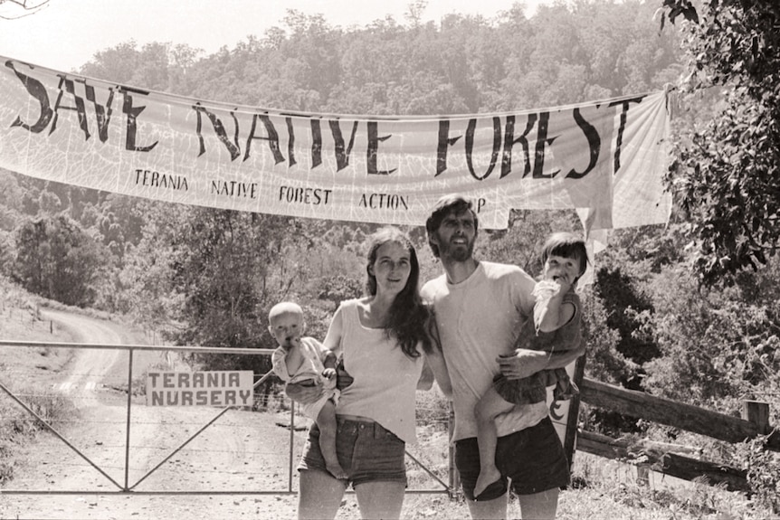 Photo from the 1970s of a young couple with small children on their hips in front of a banner that says 'save native forest'.