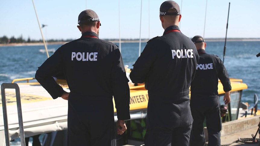 Three Water Police officers dressed in blue uniforms with Police written on the back stand in front of a boat.