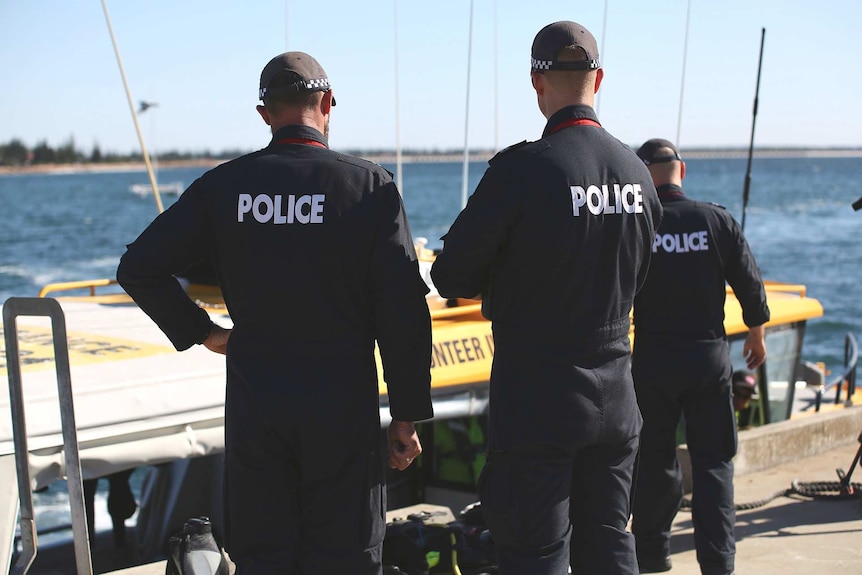 Three Water Police officers dressed in blue uniforms with Police written on the back stand in front of a boat.