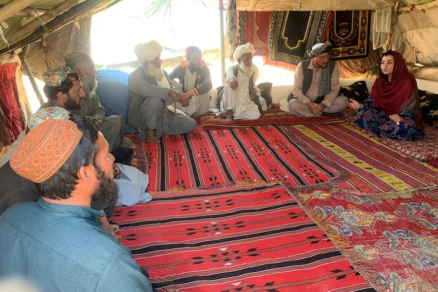 A woman speaks to a group of men from inside a tent.