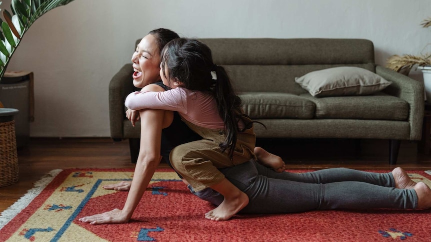 A woman doing yoga in a loungeroom with a young child climbing on her.