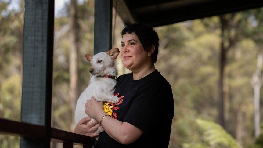 A white woman with short hair holds a white dog on a verandah