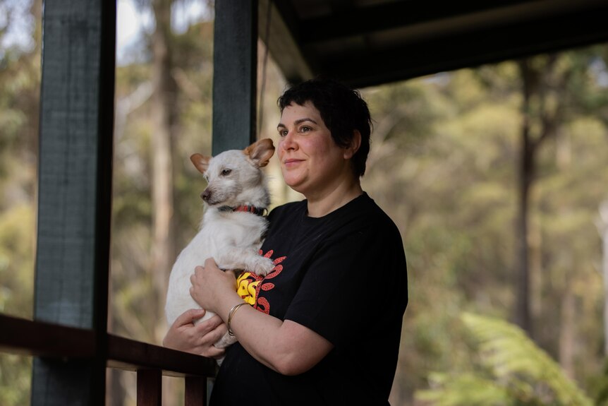 A white woman with short hair holds a white dog on a verandah
