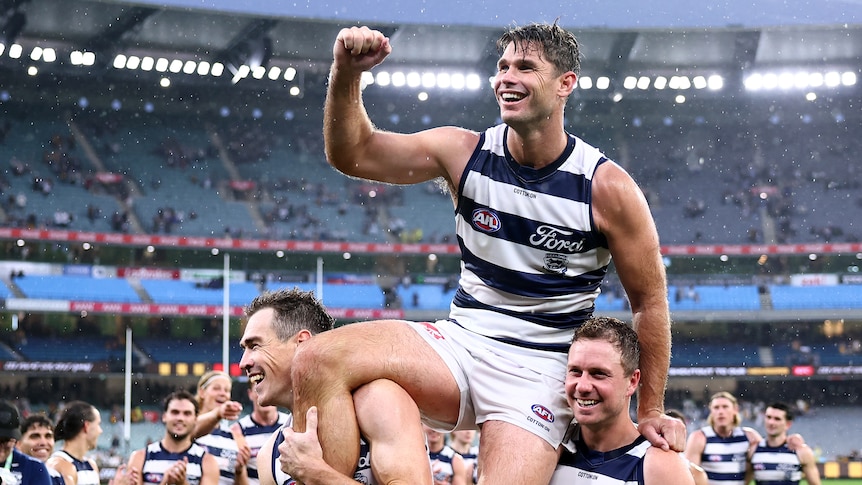Tom Hawkins is chaired off the MCG field by Geelong Cats teammates Jeremy Cameron and Mitch Duncan in the rain.