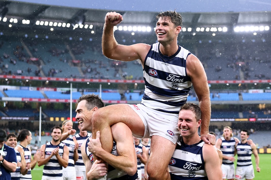 Tom Hawkins is chaired off the MCG field by Geelong Cats teammates Jeremy Cameron and Mitch Duncan in the rain.