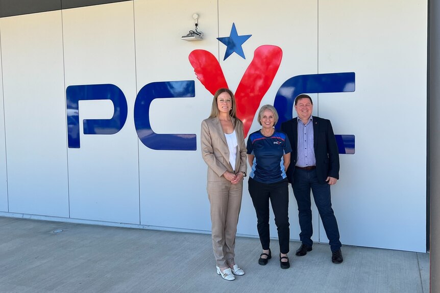 Two women and a man stand side by side in front of a building, with a sign that reads "PCYC"