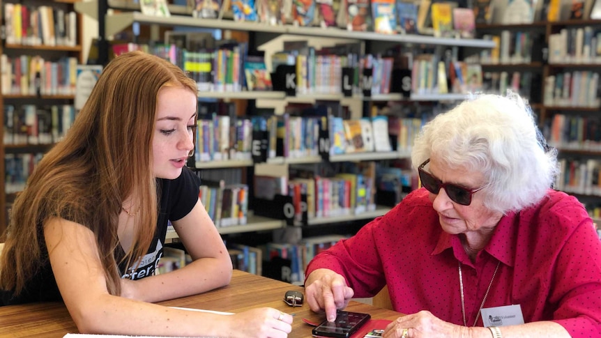 A teenage girl sits with elderly woman Ruth Holmes and teaches her how to use some apps on her phone
