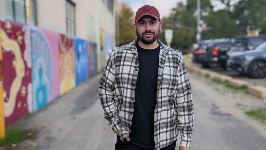 Darcie stands in a hat and plaid shirt in the foreground in the centre of an alley with a mural to his left