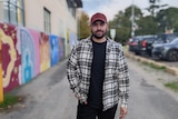 Darcie stands in a hat and plaid shirt in the foreground in the centre of an alley with a mural to his left