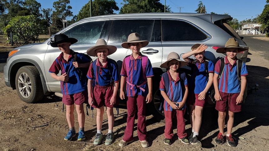 Six kids stand in front of silver SUV car wearing school uniforms, backpacks and broad brimmed hats