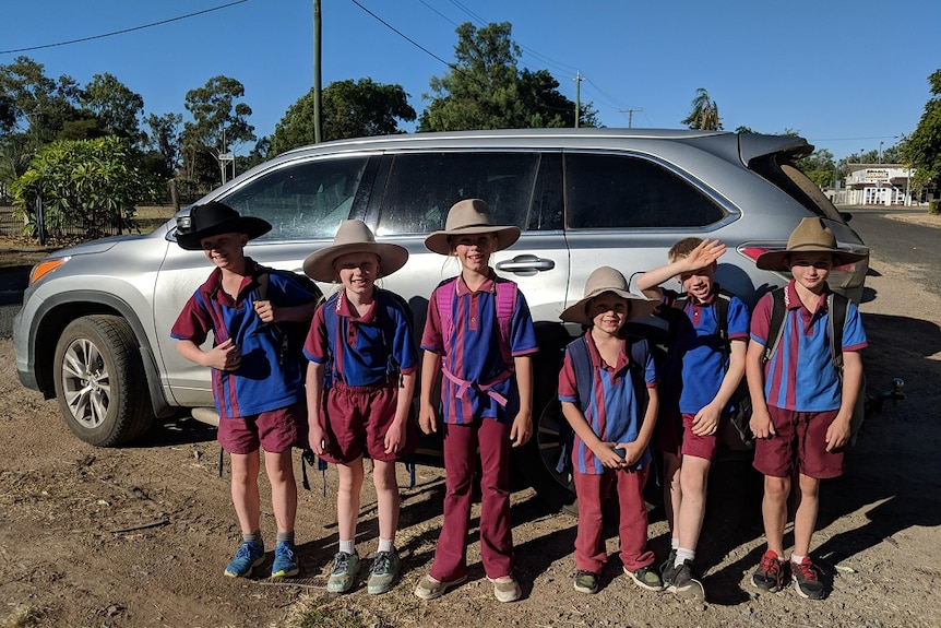 Six kids stand in front of silver SUV car wearing school uniforms, backpacks and broad brimmed hats
